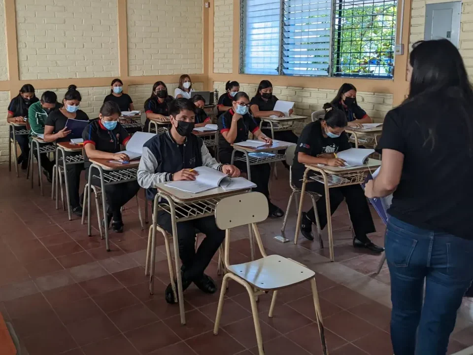 Students in face masks at their desks, workbooks open, while a teacher stands in front of the class