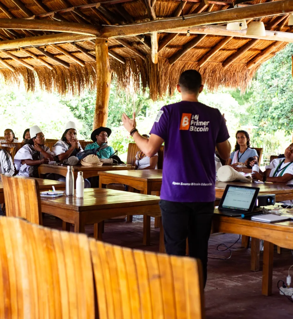 An educator in a Mi Primer Bitcoin T-shirt teaches a class seated at wooden tables under a thatched roof